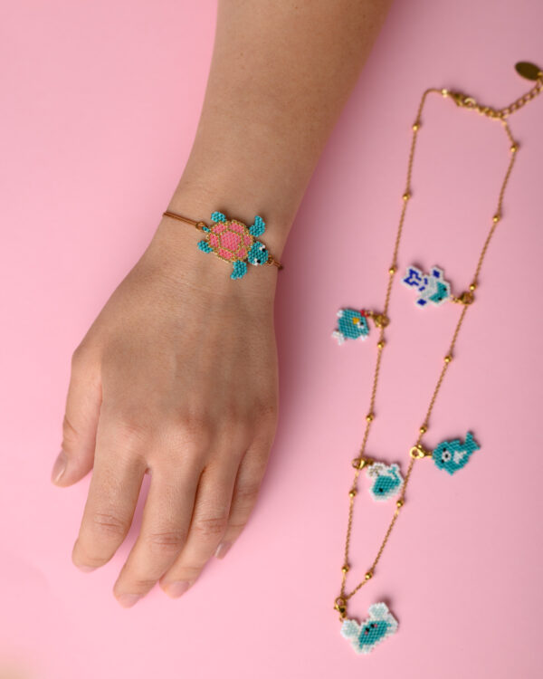 Hand displaying Miyuki sea turtle charm bracelet by The Gem Stories on a pink background.