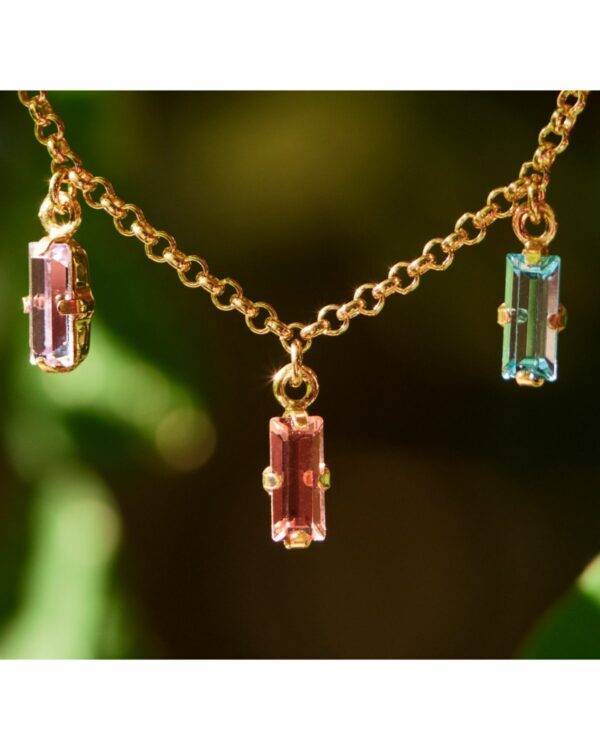 Close-up of Navete Multicolor Crystals Necklace with pink and teal crystals on a golden chain, set against a lush green background