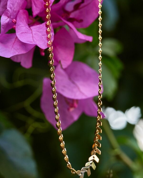 Gold necklace with a leaf design hanging against a backdrop of purple flowers