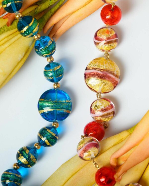 Close-up of two Murano necklaces with vibrant blue and red beads featuring gold accents, displayed on a decorative surface