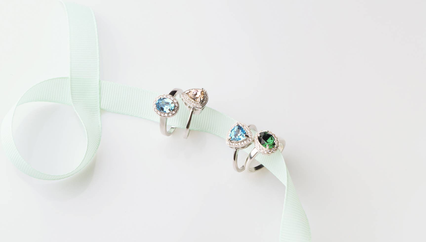 Four gemstone rings with blue, green, and peach stones surrounded by diamonds, displayed on a light green ribbon