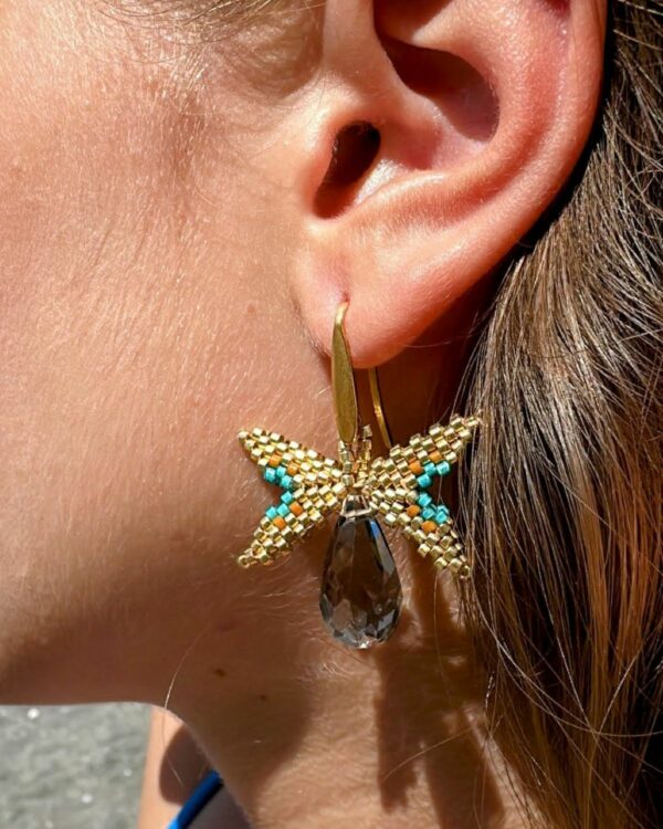 Miyuki butterfly earring with gold accents and a teardrop crystal pendant by The Gem Stories, displayed against a beach background.