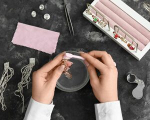 Hands cleaning jewelry with a brush, surrounded by sparkling earrings and rings in a jewelry tray, alongside cleaning tools.