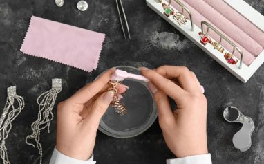 Hands cleaning jewelry with a brush, surrounded by sparkling earrings and rings in a jewelry tray, alongside cleaning tools.